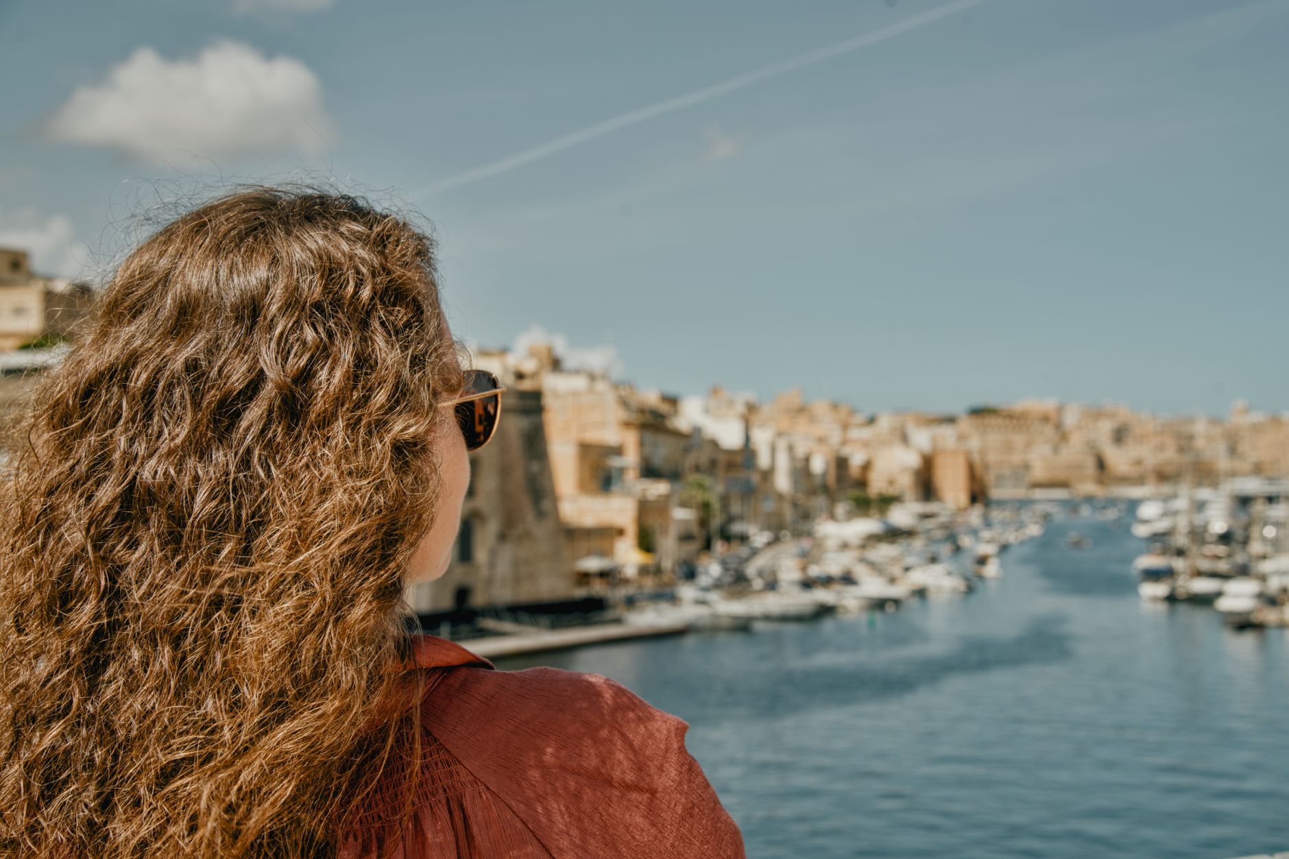 A young woman with curly hair admires the scenic marina view in Malta's historic city under clear blue skies.