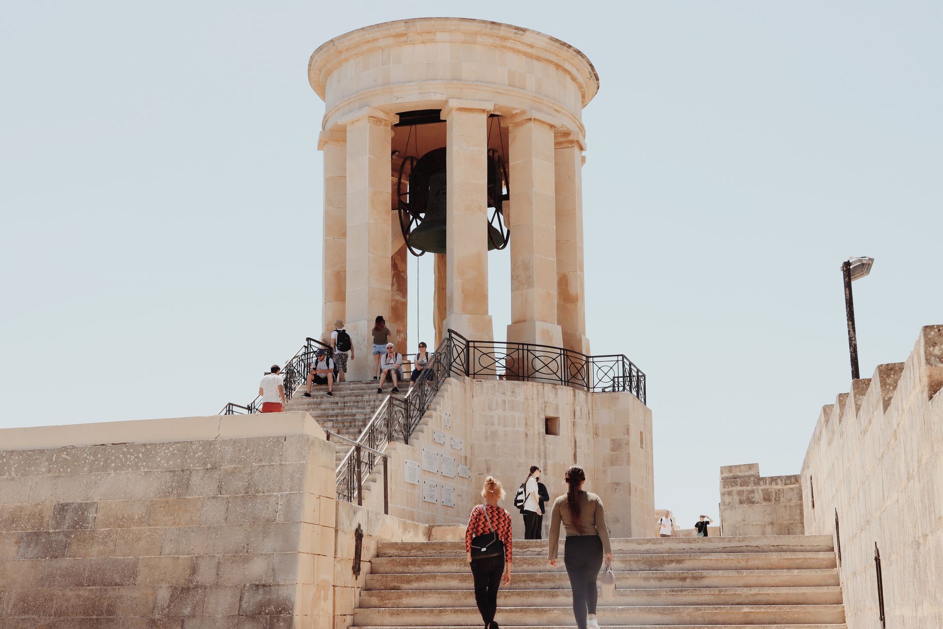 Tourists explore the iconic Siege Bell War Memorial in Valletta, Malta, on a sunny day.